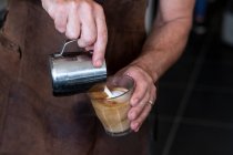 Primer plano de barista vistiendo delantal marrón vertiendo café con leche. - foto de stock