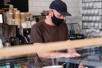 Varón barista con gorra de béisbol negra y máscara facial trabajando detrás del mostrador. - foto de stock