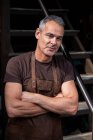 Retrato de barista masculino con pelo gris corto, con delantal marrón, brazos cruzados, mirando a la cámara. - foto de stock