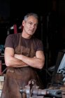 Portrait of male barista with short grey hair, wearing brown apron, arms folded, looking at camera. — Fotografia de Stock