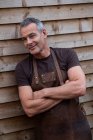 Portrait of male barista with short grey hair, wearing brown apron, arms folded, leaning against wooden wall. — Stock Photo