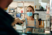 Woman in a face mask behind cafe counter with safety screen, offering a contactless payment terminal to a customer — Stock Photo