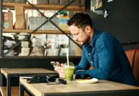 Человек, сидящий один за столом с ноутбуком и соком напиток в кафе с помощью смартфона — стоковое фото