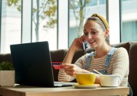 Mulher sentada em um café usando um laptop e falando em um telefone inteligente, segurando um cartão de crédito — Fotografia de Stock