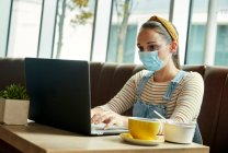 Femme portant un masque facial assis dans un café à l'aide d'un ordinateur portable — Photo de stock