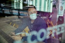 Homem com uma máscara facial sentado em uma mesa de café, vista através de uma janela — Fotografia de Stock