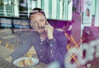 Uomo con una maschera facciale seduto a un tavolo da caffè con un telefono cellulare, vista attraverso una finestra — Foto stock
