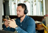 Hombre sentado en un café usando auriculares, usando un teléfono inteligente, trabajando remotamente. - foto de stock