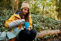 Frau sitzt auf Baumstamm im Wald und gießt Getränk aus Thermoskanne — Stockfoto