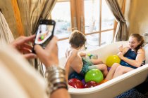 Person taking a smart phone photograph of a young boy and his older sister in bathtub with water balloons — Stock Photo