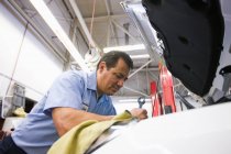 Mecánico hispano se inclina en un motor de un coche en el que trabaja en un taller de reparación de automóviles - foto de stock