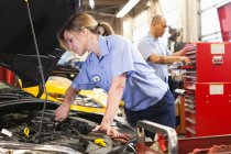 Caucasian female mechanic working on engine in auto repair shop with Hispanic coworker in background — Stock Photo