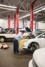 Mecânico hispânico apoiado em um carro como ele trabalhando no compartimento do motor em uma oficina de reparação de automóveis — Fotografia de Stock