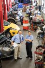 Retrato del propietario del taller de reparación de automóviles hablando con la mecánica femenina como colegas que trabajan en segundo plano - foto de stock