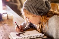 Adolescente dans un dessin de chapeau laineux avec un crayon sur un bloc-croquis. — Photo de stock