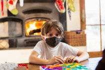 Jovem menino usando máscara jogando jogo de tabuleiro em casa — Fotografia de Stock