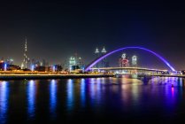 Notte, paesaggio urbano di Dubai, edifici alti e lungomare, e arco sul torrente. — Foto stock