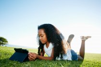 Nove anni mista ragazza di razza sdraiata sull'erba guardando un tablet digitale. — Foto stock