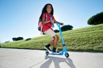 Young mixed race girl in pink shirt and formal tie, wearing a backpack on a scooter — Stock Photo