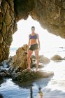 Молода жінка, тріатлоністка в тренуванні в капелюсі для плавання та окулярах, переглянута через кам'яну арку на пляжі — стокове фото