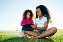 Jovem menina de raça mista e sua irmã mais nova sentada ao ar livre compartilhando um tablet digital — Fotografia de Stock