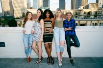 Five young women posing for photographs on a rooftop, city skyline background — Stock Photo