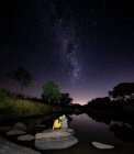 Man and woman sit on a boulder together next to a river watching the milkway — Stock Photo