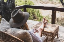 Woman sitting with her feet up drinking a cup of tea, wearing a safari hat — Stock Photo