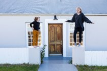 Hermano y hermana de pie en una pared baja fuera de una casa riendo - foto de stock