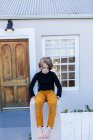 Мальчик, сидящий на стене возле своего дома, ждет или ему скучно — стоковое фото