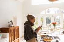 Mulher adulta na cozinha ler texto no telefone inteligente e cozinhar uma refeição. — Fotografia de Stock