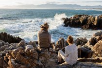 Woman and teenage girl sitting on rocky shore, looking out to sea. — Stock Photo