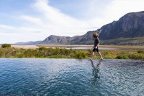 Восьмилетний мальчик прогуливается по краю бесконечного бассейна, на фоне гор — стоковое фото