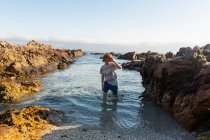 A young boy in shallow sea water among jagged rocks on the beach at De Kelders. — Stock Photo