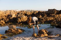 Niño explorando una piscina de rocas entre las rocas dentadas de la costa del Océano Atlántico al atardecer, De Kelders, Cabo Occidental, Sudáfrica. - foto de stock