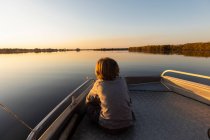 Jeune garçon assis à l'arrière d'un bateau sur le delta de l'Okavango au coucher du soleil — Photo de stock