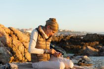 Mulher adulta sentada na praia usando seu smartphone no De Kelders ao pôr do sol. — Fotografia de Stock