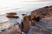 Una familia, madre y dos niños sentados mirando el atardecer sobre el océano. - foto de stock