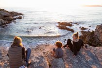 Une famille, une mère et deux enfants assis à regarder le coucher du soleil sur l'océan. — Photo de stock