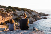 Adolescente y joven sentado en las rocas mirando sobre el mar al atardecer - foto de stock