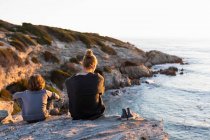 Adolescente y joven sentado en las rocas mirando sobre el mar al atardecer - foto de stock