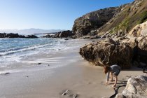 Un giovane ragazzo solo su una piccola distesa di sabbia sotto le scogliere vicino all'oceano. — Foto stock