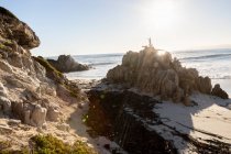 Um menino em pé em cima de uma rocha acima de uma praia arenosa — Fotografia de Stock