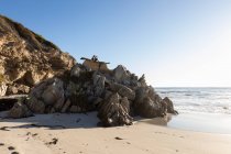 Duas crianças empoleiradas em cima de rochas irregulares com vista para uma praia de areia na maré baixa — Fotografia de Stock