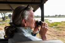 Donna anziana con binocolo, seduta in un veicolo safari, che guarda oltre paludi e corsi d'acqua — Foto stock