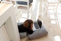 Achtjähriger Junge liegt auf Kissen, Kinn auf Händen, schaut auf Laptop-Bildschirm, macht Hausaufgaben. — Stockfoto