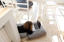 Восьмилетний мальчик лежит на подушках, подбородок на руках, смотрит на экран ноутбука, делает домашнюю работу. — стоковое фото