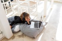 Otto anni ragazzo sdraiato sul pavimento su cuscini utilizzando un computer portatile — Foto stock