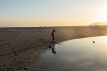 Niño en una playa, mirando su reflejo en una piscina de agua, marea baja, puesta de sol. - foto de stock