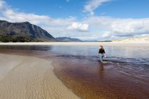 Una adolescente vadeando a través de un canal de agua en una amplia playa de arena. - foto de stock